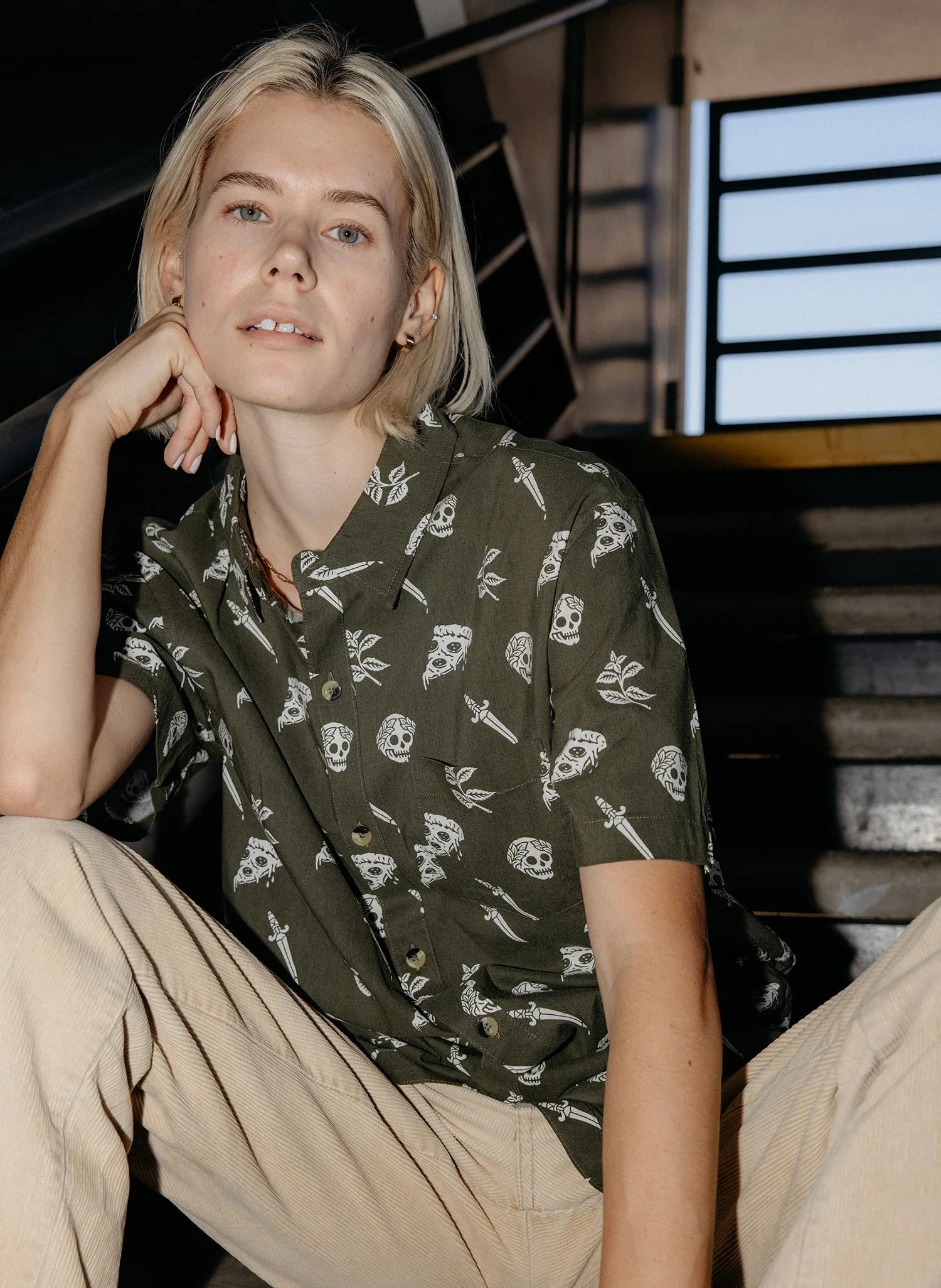 Person in olive green button-up shirt with white skull and dagger pattern, sitting on stairs