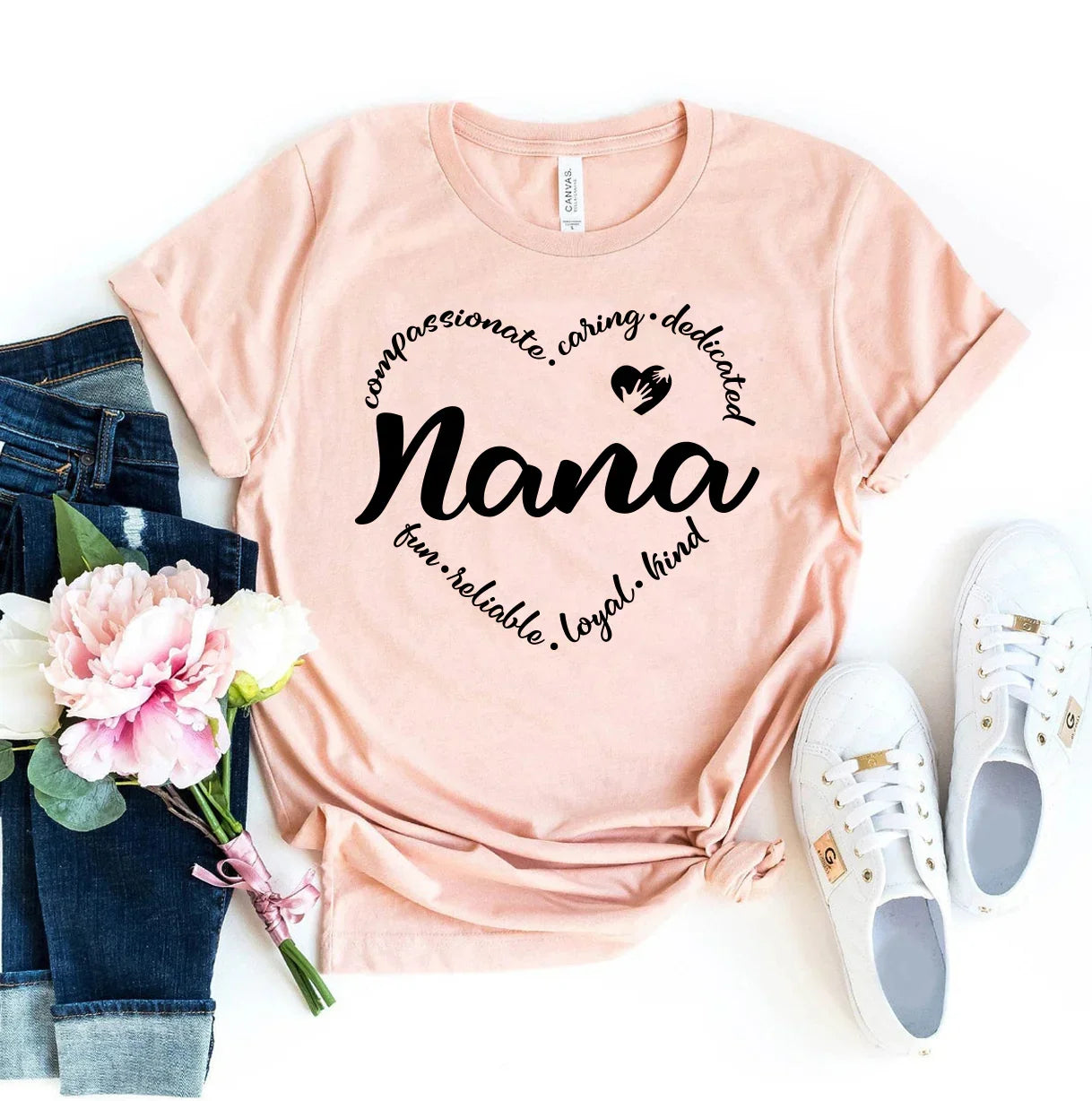 Light pink t-shirt with black text Nana surrounded by caring words, paired with jeans, white sneakers, and pink flowers