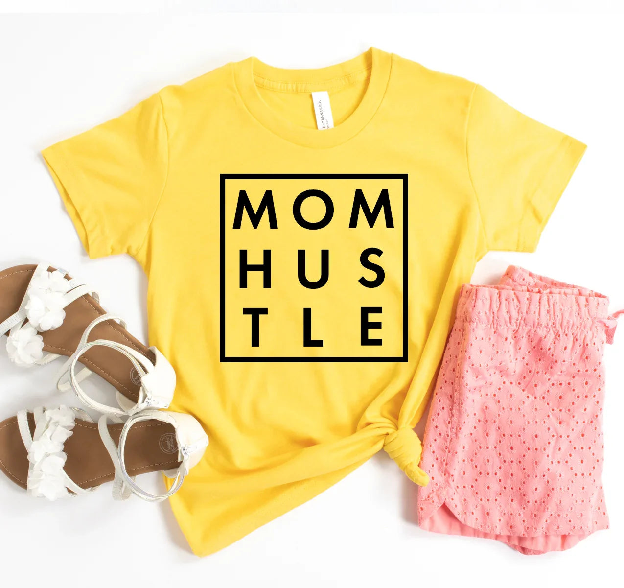 Yellow t-shirt with black MOM HUSTLE text, pink eyelet shorts, and white sandals flat lay