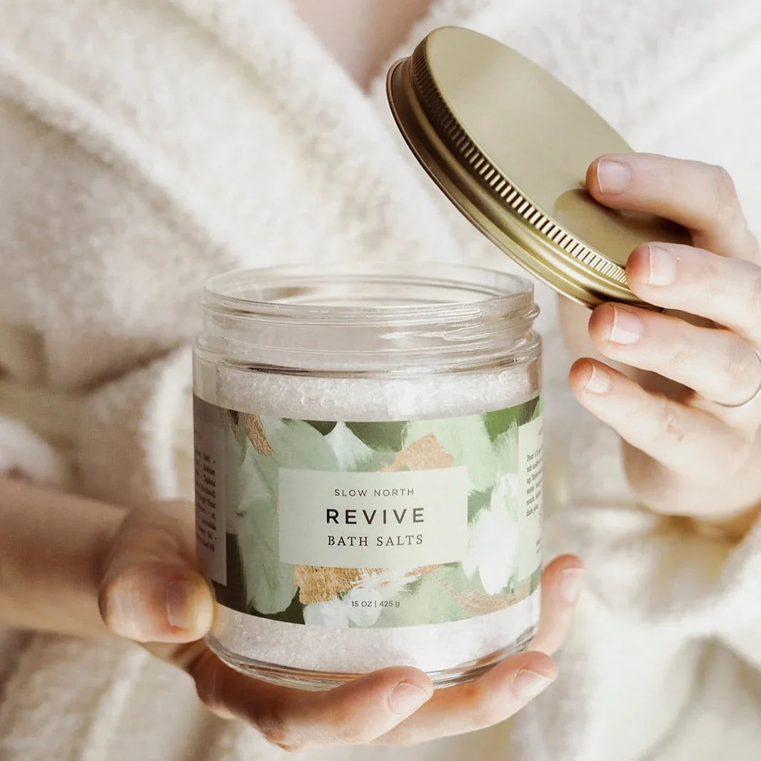 Person holding open jar of Slow North Revive bath salts with floral label and gold lid