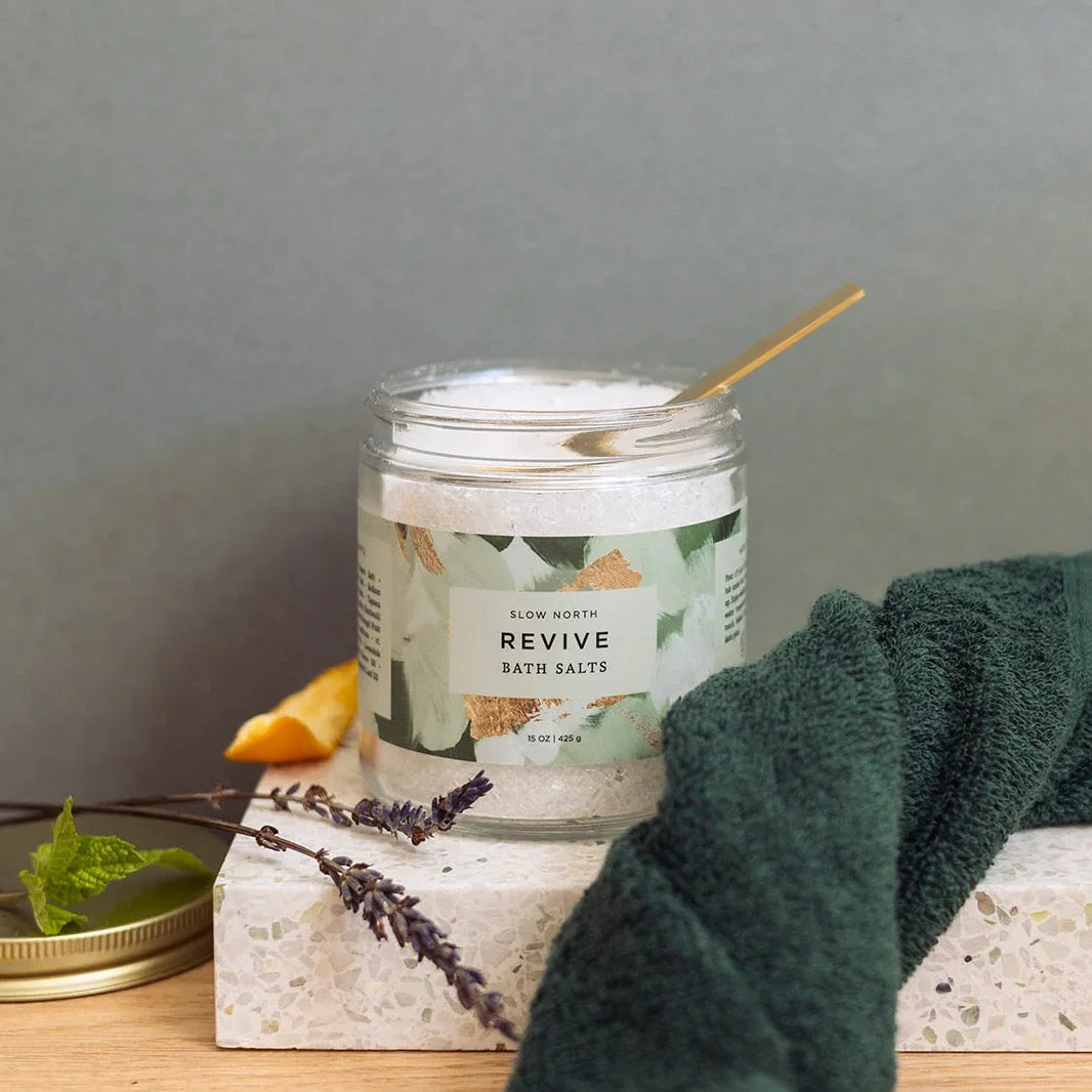 Jar of Slow North Revive bath salts with a spoon, lavender sprigs, lemon peel, and green towel on terrazzo surface