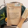 Slow North Revive bath salts pouch with natural ingredients, bath brush, and green towel in a cozy spa setting at Qu!rk Boutique.