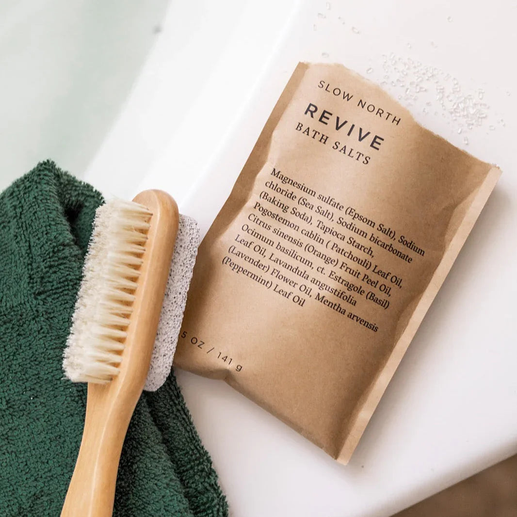 Slow North Revive bath salts pouch on a bath ledge with brush and green towel, Qu!rk Boutique