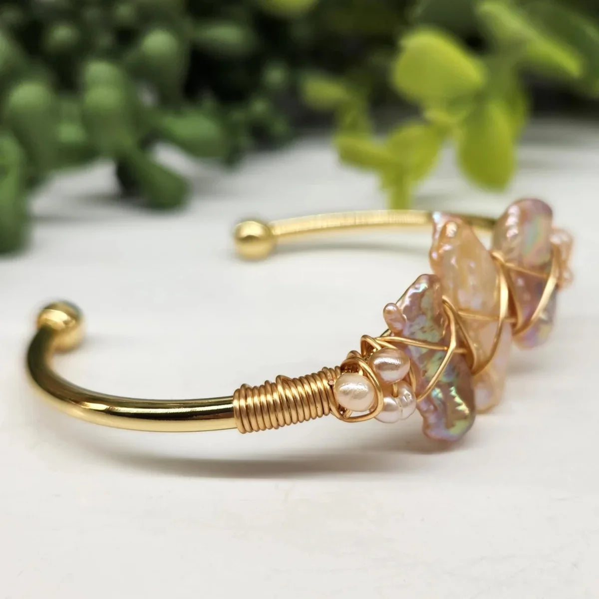 Gold wire-wrapped bangle bracelet with iridescent stones and pearls on white, green plant background
