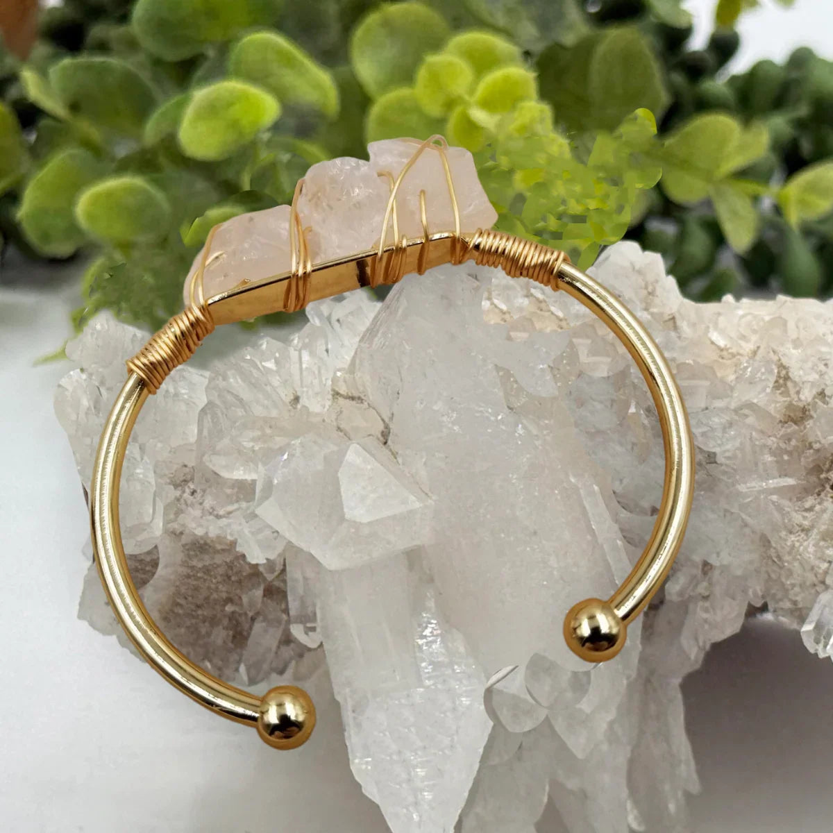 Gold cuff bracelet with raw pink crystals, displayed on clear quartz, plants in background