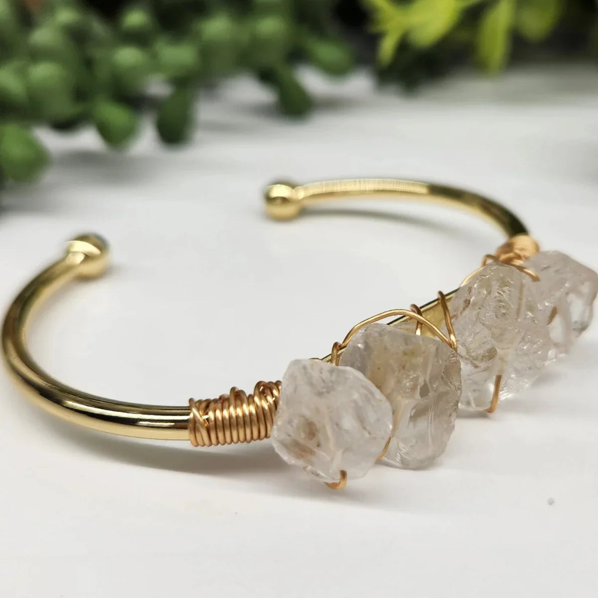 Gold wire cuff bracelet with raw clear crystal stones, displayed on white, greenery in background