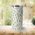 White travel tumbler with green botanical leaf pattern on wooden table with blurred natural background