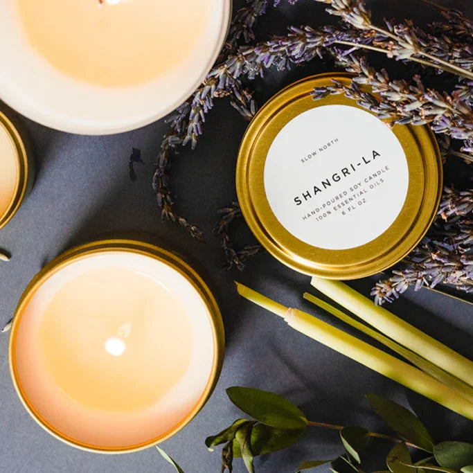 Shangri-La 6oz hand-poured soy candle in gold tin, surrounded by lavender and lemongrass, at Qu!rk Boutique.