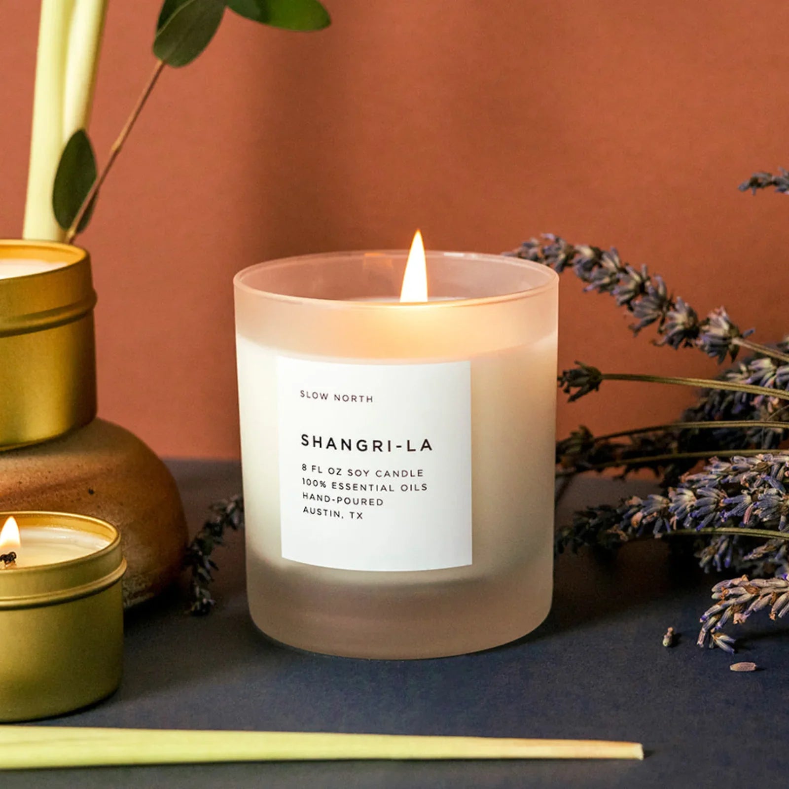 Shangri-La soy candle in frosted glass, lit and surrounded by lavender and gold tins