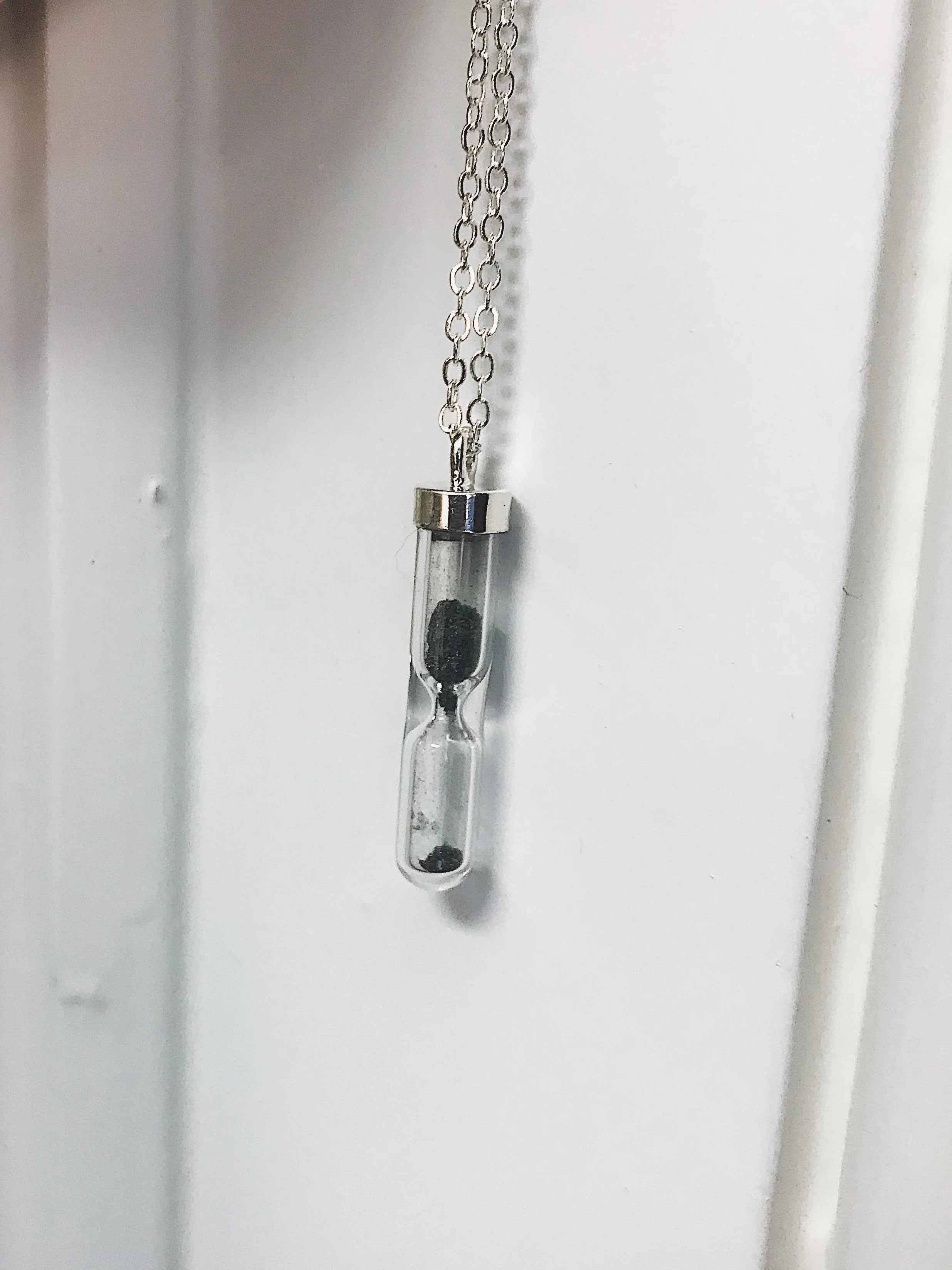 Silver chain necklace with a small glass hourglass pendant containing black sand, hanging against a white background