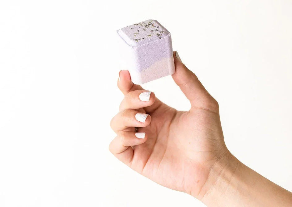 Hand holding a lavender and white square shower steamer with dried flowers on top