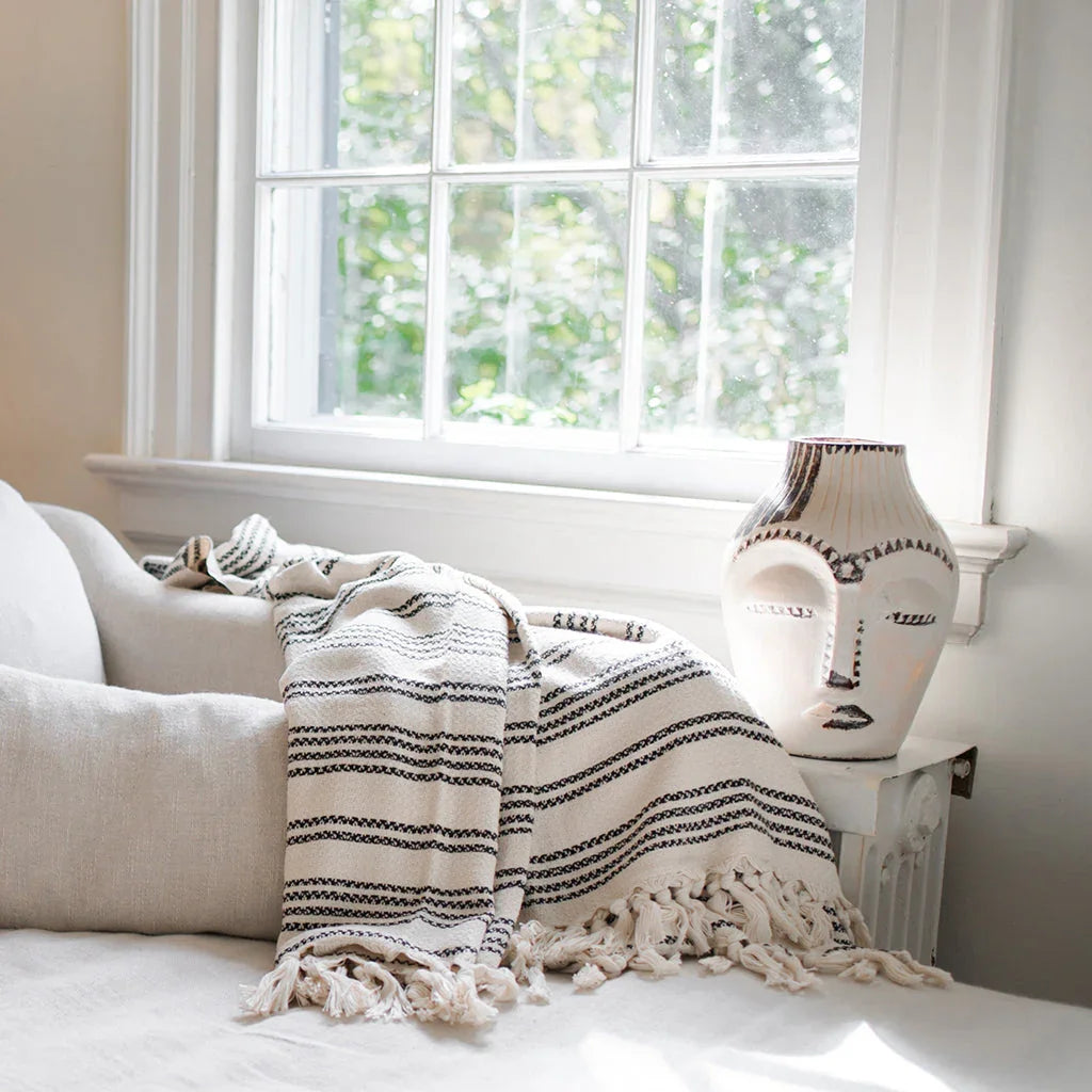 Woven striped throw blanket with fringe on a sofa beside a decorative vase in a sunlit room