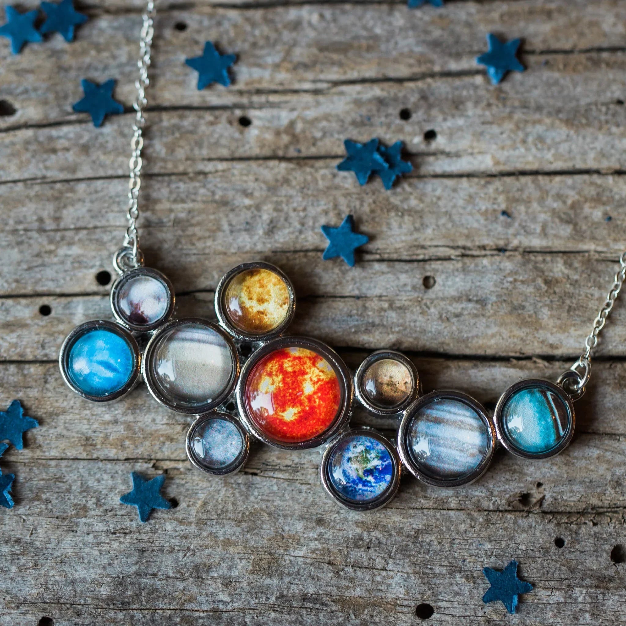 Silver necklace with circular pendants featuring colorful planet designs on rustic wooden background with blue star confetti
