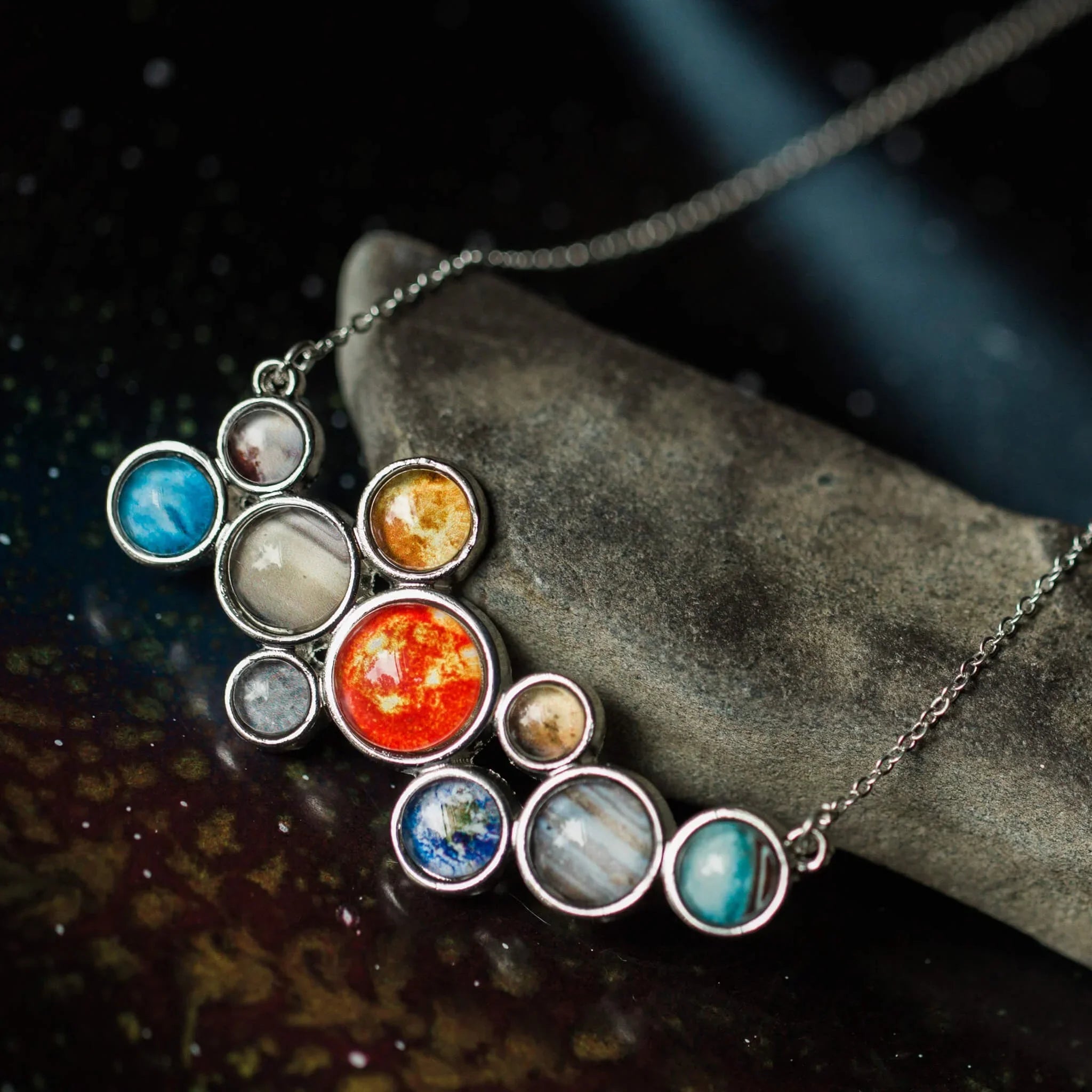 Silver planetary necklace with colorful planet designs on a black and gray textured background