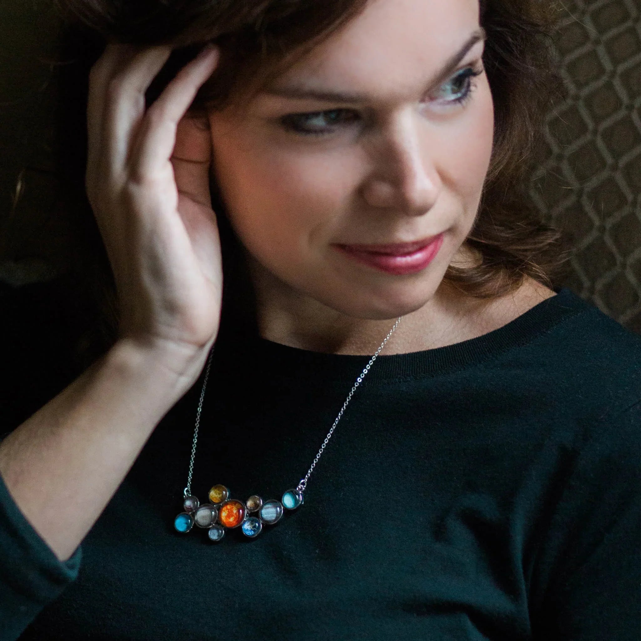 Woman wearing a black top and solar system pendant necklace, hand touching her hair