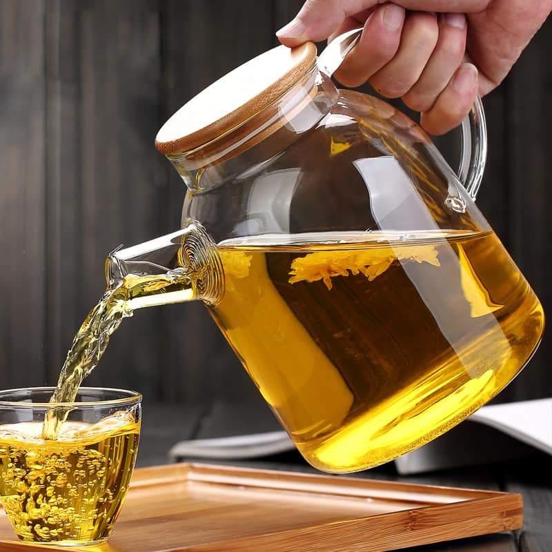 Glass teapot with wooden lid pouring herbal tea into a cup on a wooden tray
