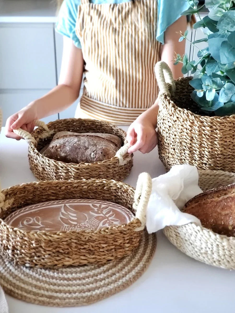 Person in striped apron holding woven bread basket with dark bread on white table with other baskets and plant