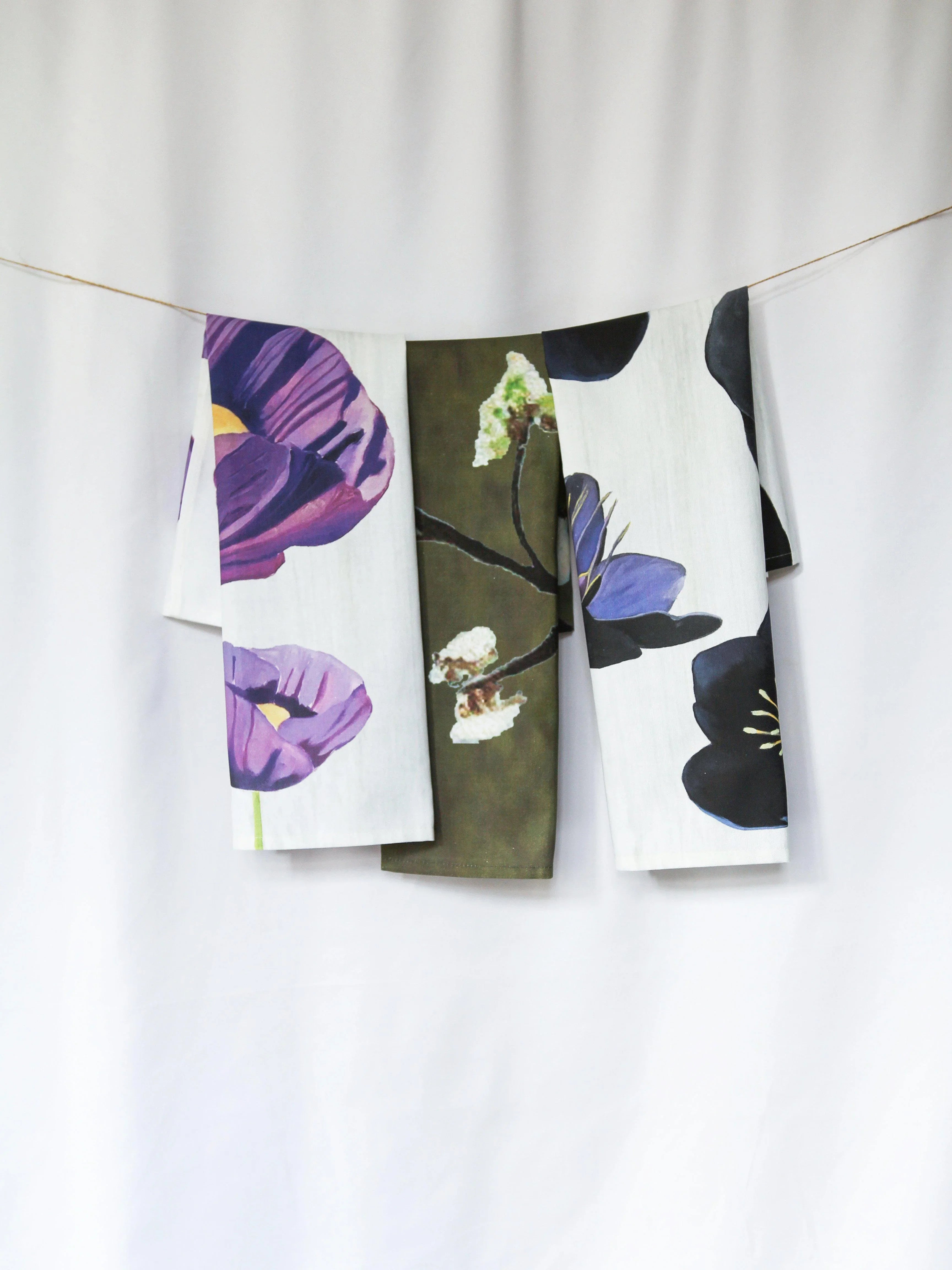 Three floral tea towels in purple, white, and olive green hanging on a string against a white background