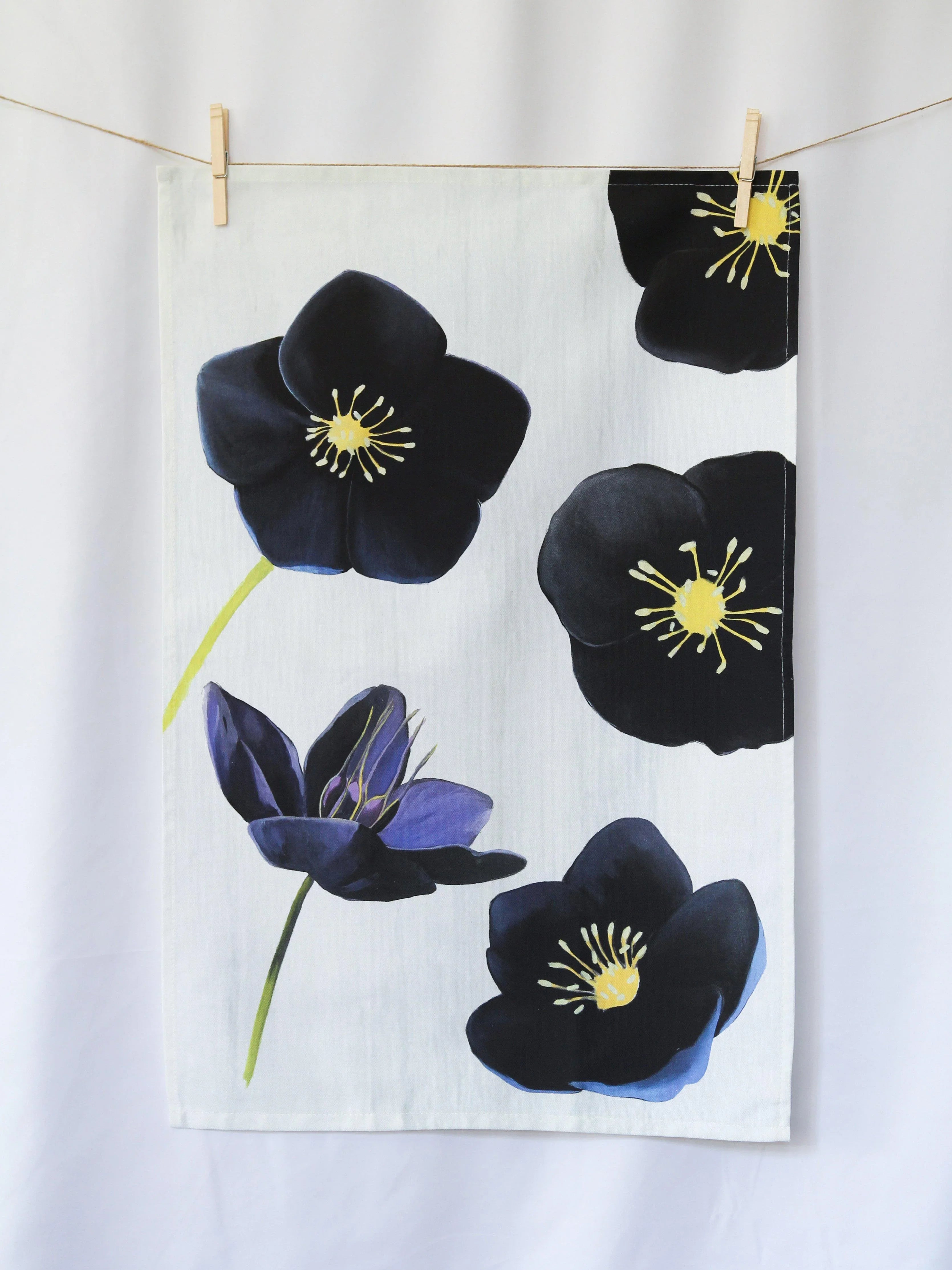 White tea towel with large black hellebore flowers and yellow centers hanging on a clothesline