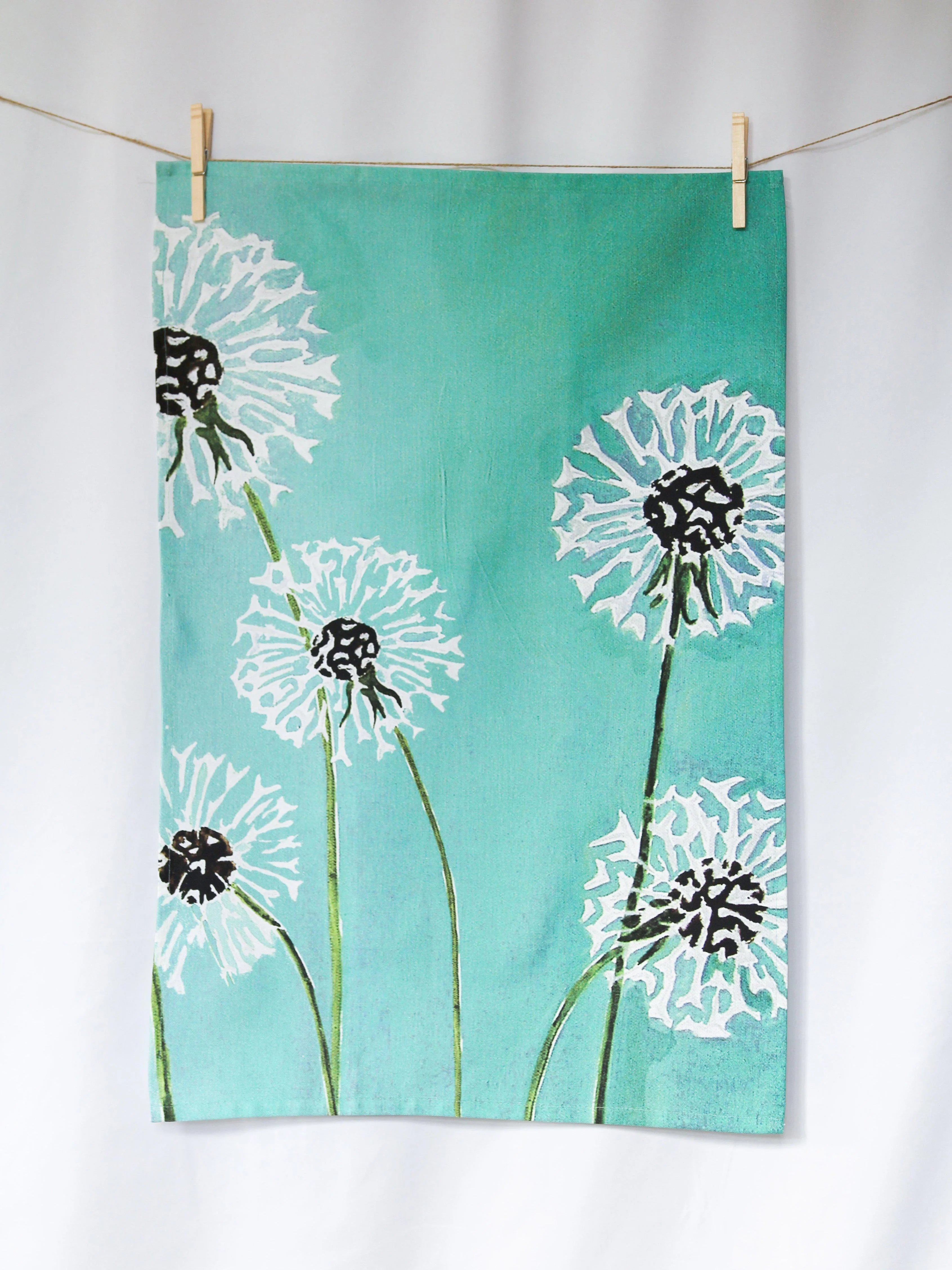Aqua tea towel with white dandelion designs and green stems hanging on a clothesline with clothespins
