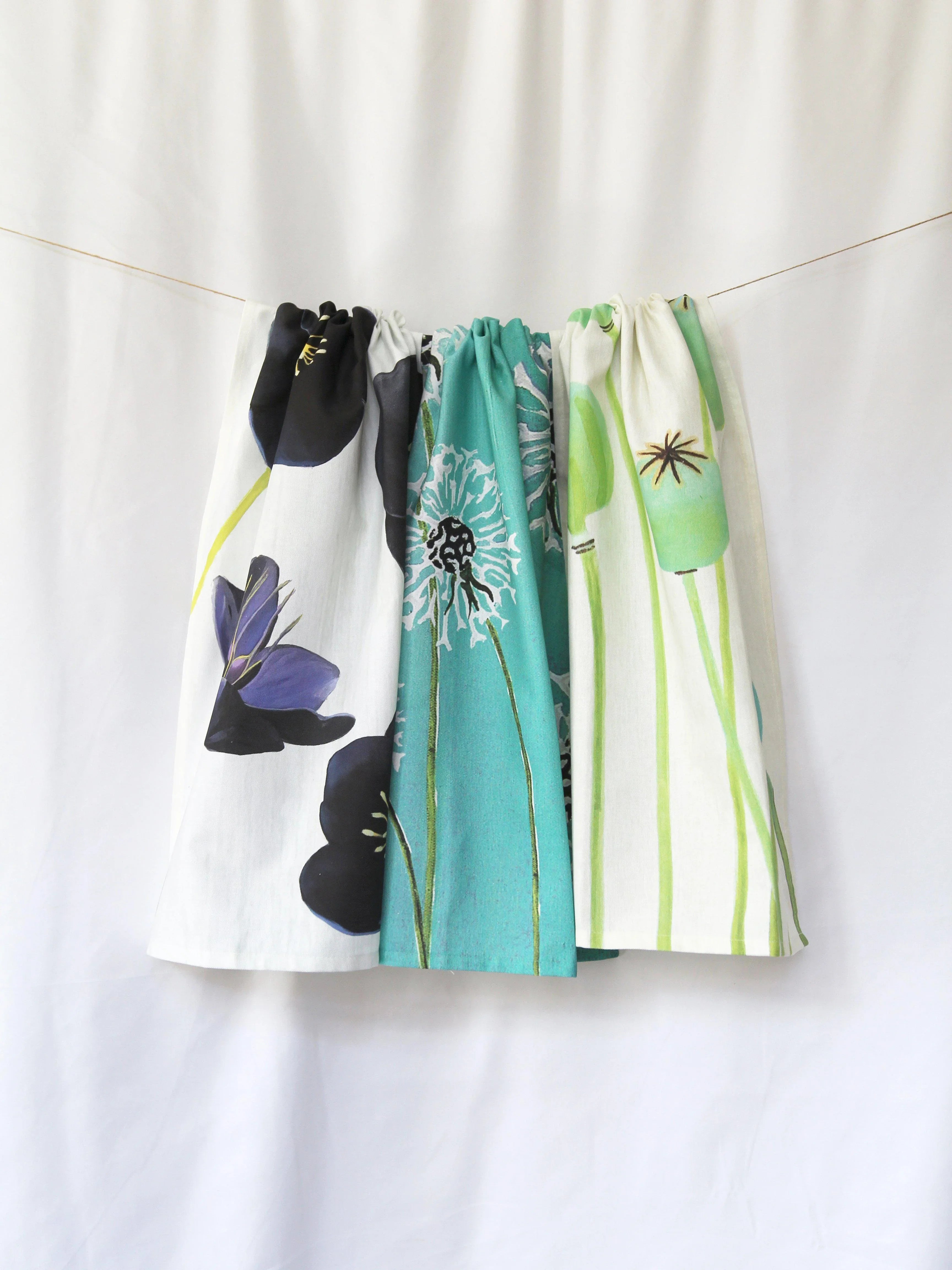Three floral tea towels with poppy pods and dandelions hanging on a string