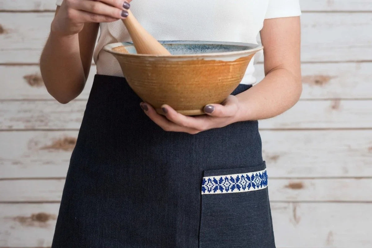 Woman wearing blue Tatreez apron with embroidered pocket, holding ceramic mixing bowl