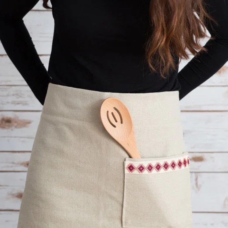 Cream apron with red embroidered pocket and wooden spoon, modeled against rustic wood backdrop.