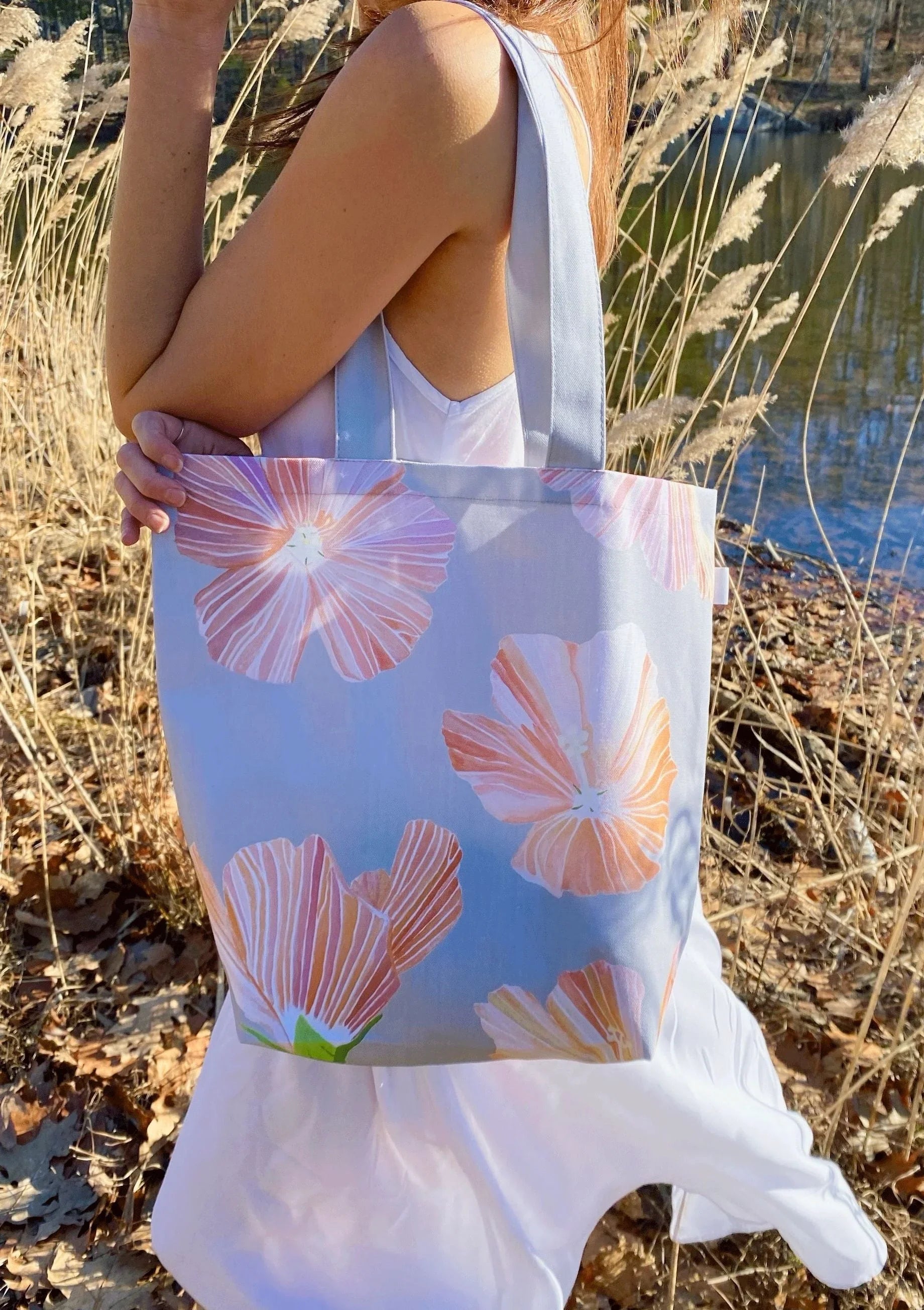 Grey tote bag with large pink hibiscus flower prints held by person in white dress near dry grass and water