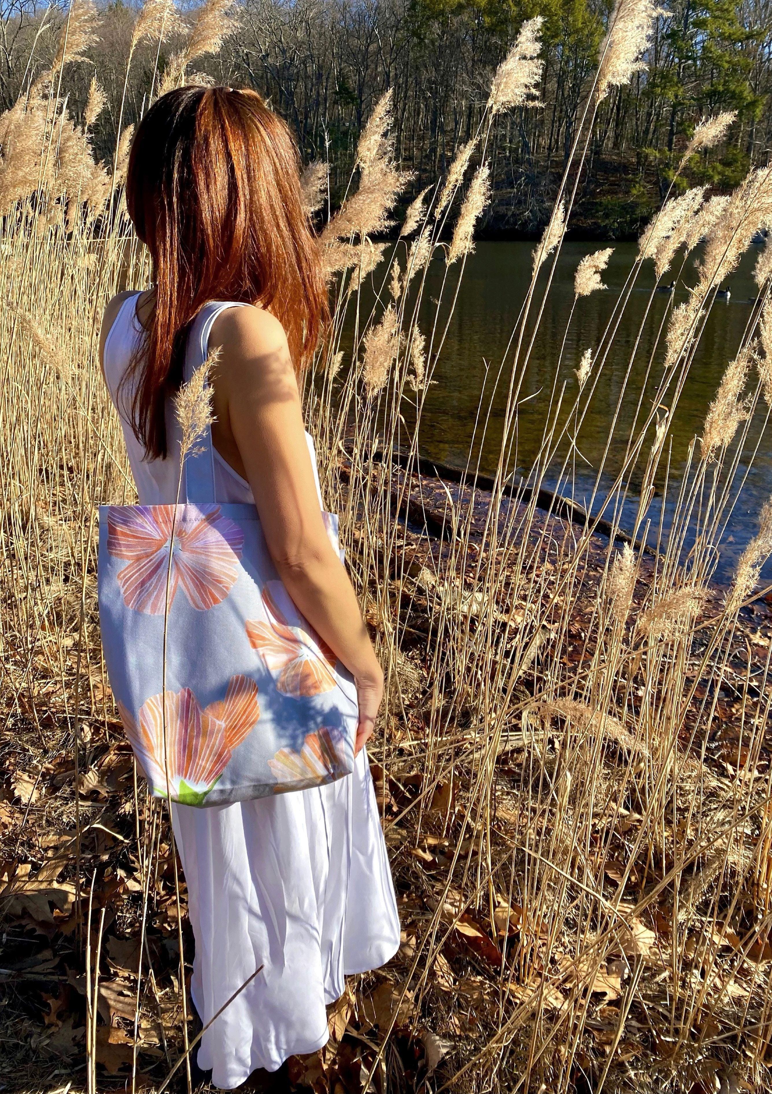 Woman in white dress standing by lake with tall dry grass holding floral pattern tote bag