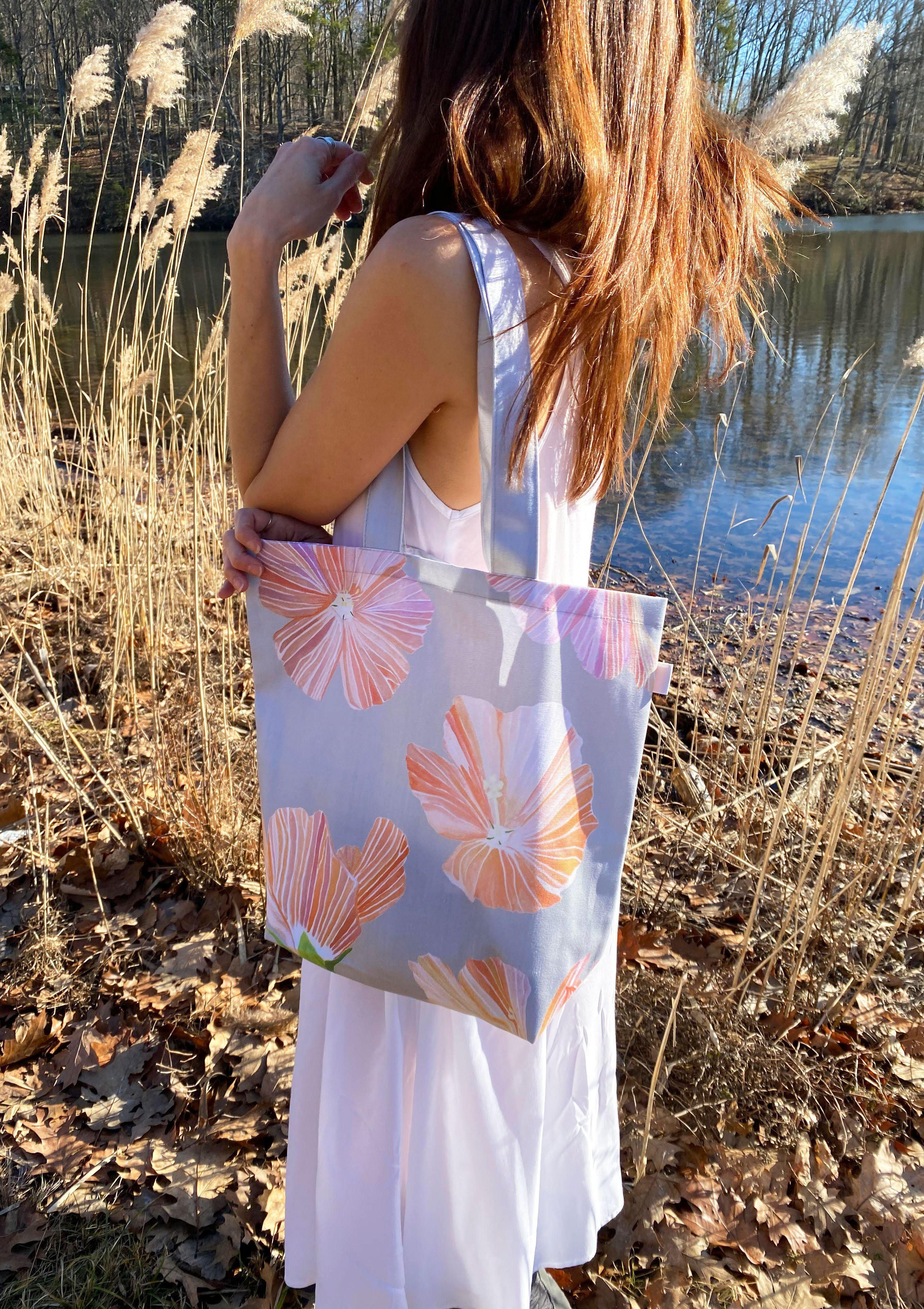 Woman wearing white dress holding tote bag with orange hibiscus floral print near lake and dry foliage