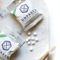 Unpaste toothpaste tabs packets and white tablets on marble surface with a bamboo toothbrush