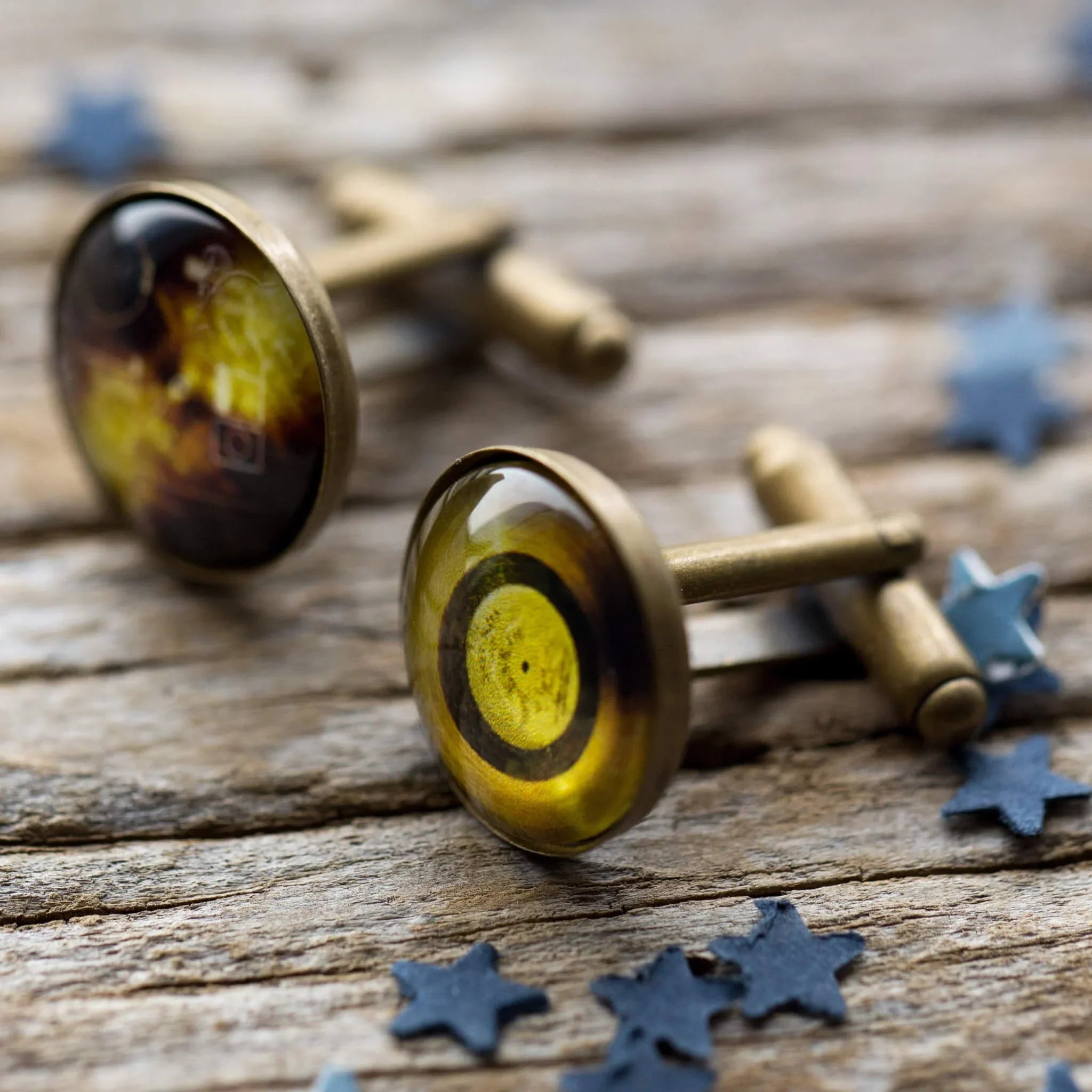 Close-up of vintage brass cufflinks with amber glass tops on rustic wooden surface with blue star confetti