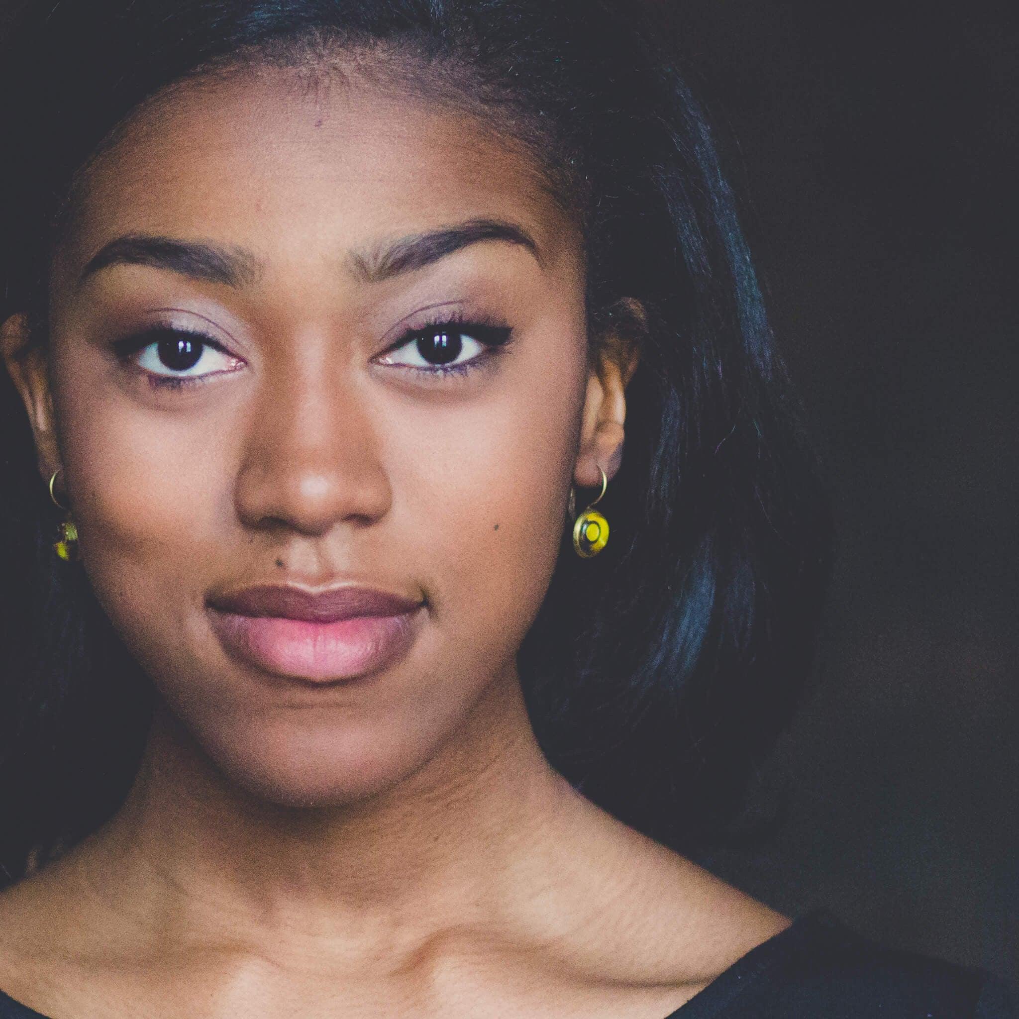 Close-up portrait of a young woman with brown skin, natural makeup, and yellow earrings against a dark background