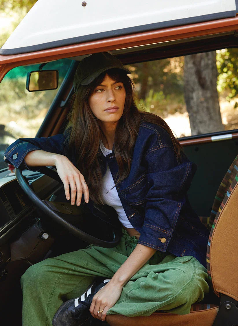 Woman in vintage washed black dad hat, denim jacket, and green pants sitting in a car