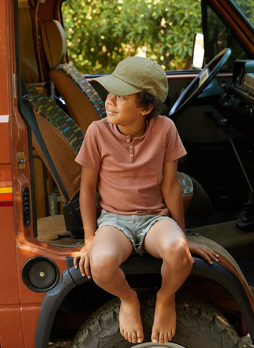Child in olive vintage washed dad hat and casual outfit sits on van tire, outdoors at Qu!rk Boutique