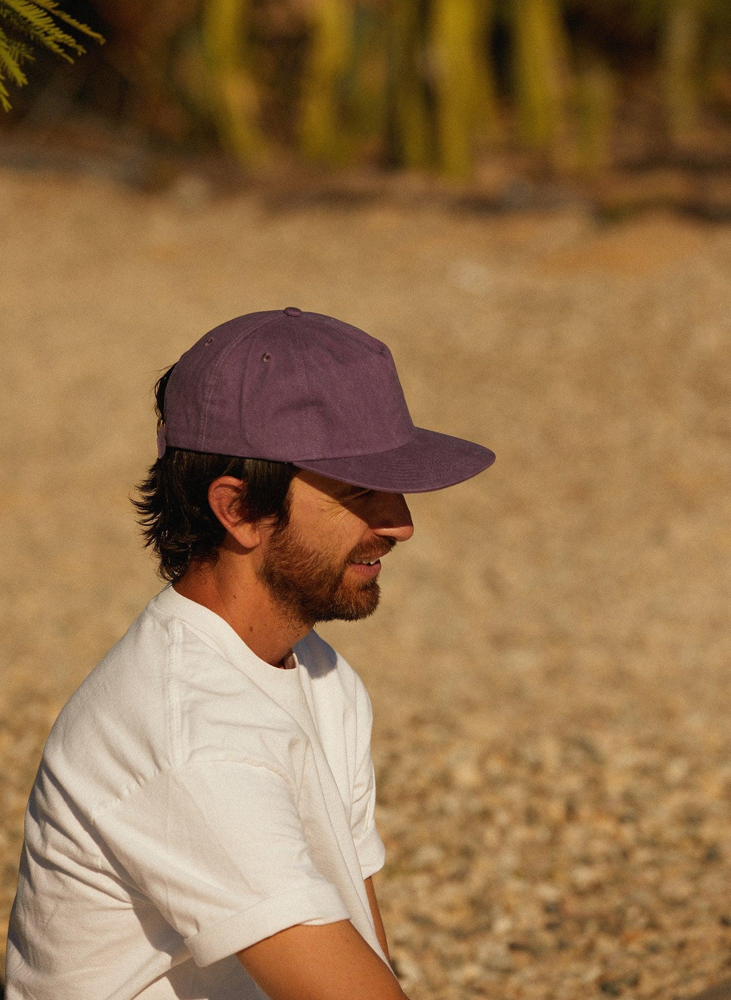 Man in a white t-shirt and purple cap outdoors on gravel, casual Qu!rk Boutique style.