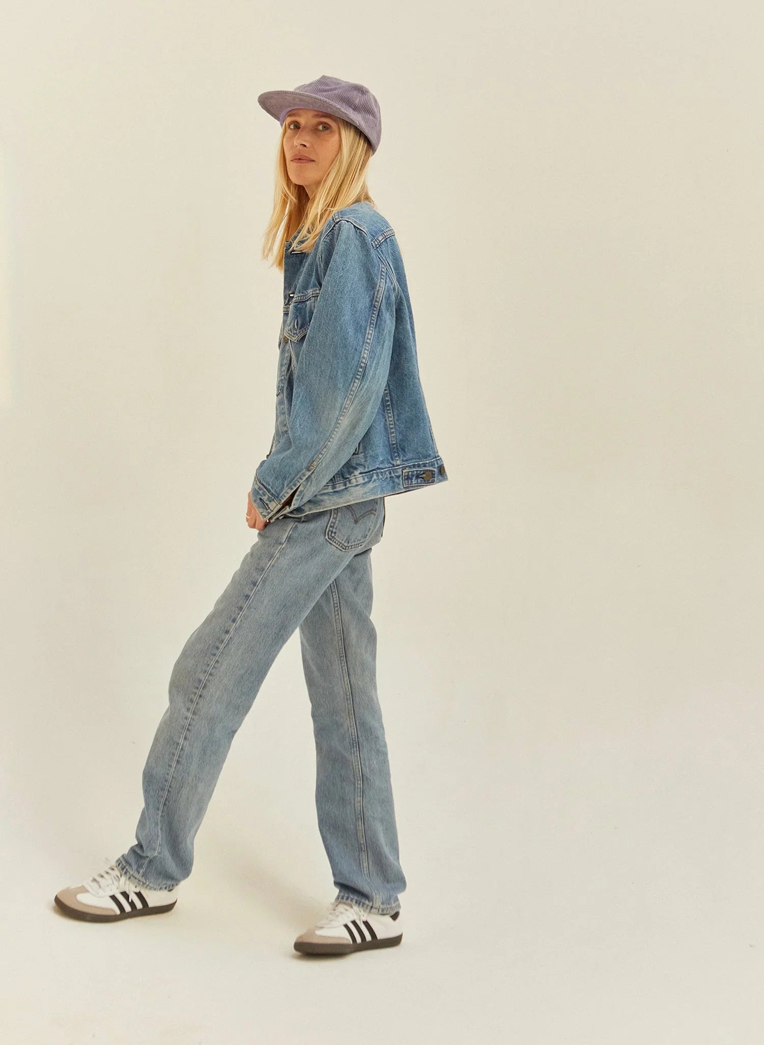 Woman in light purple corduroy hat, denim jacket, jeans, and sneakers, minimalist background