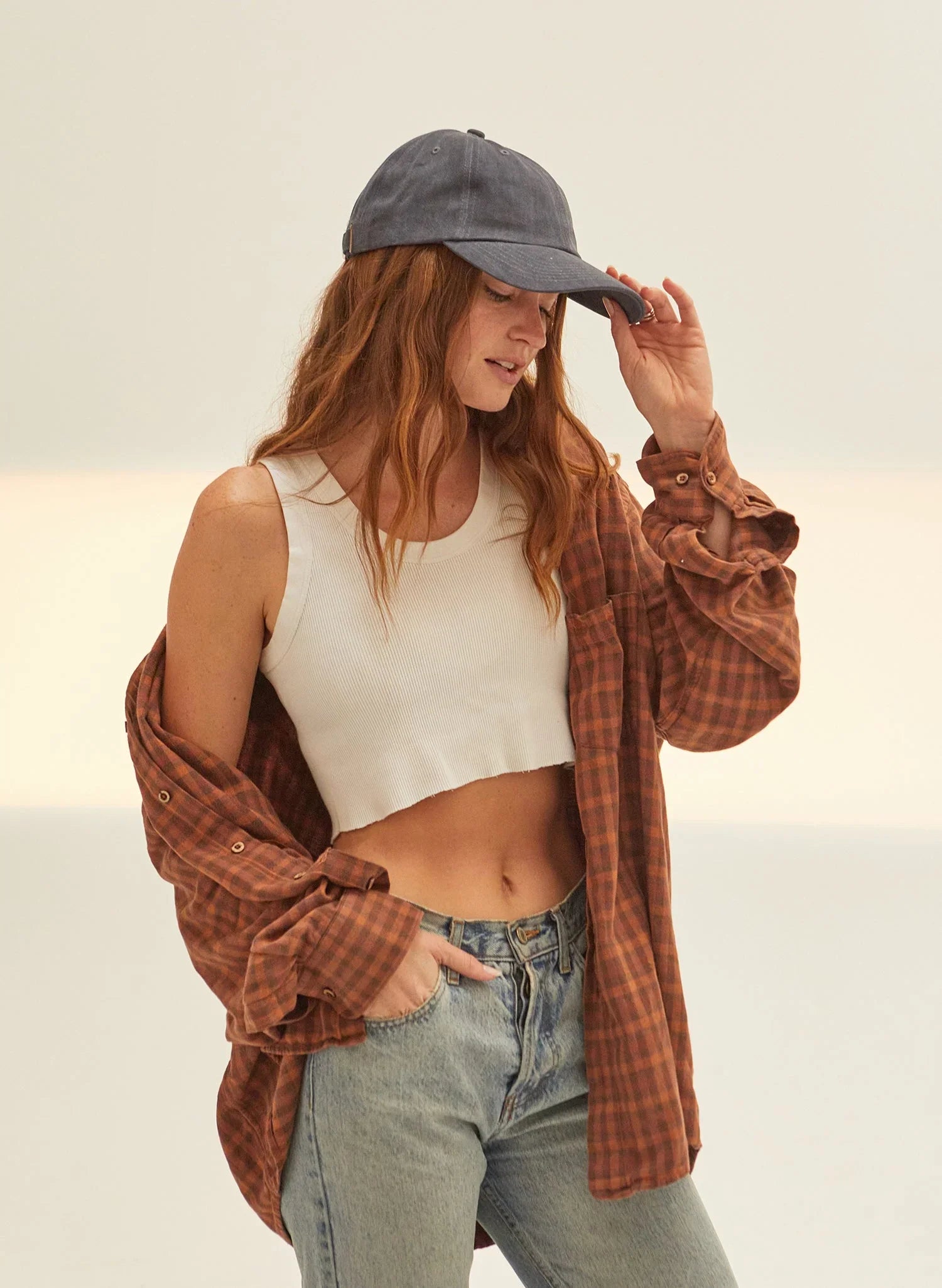 Woman wearing vintage washed black dad hat, white crop tank, plaid shirt, and jeans