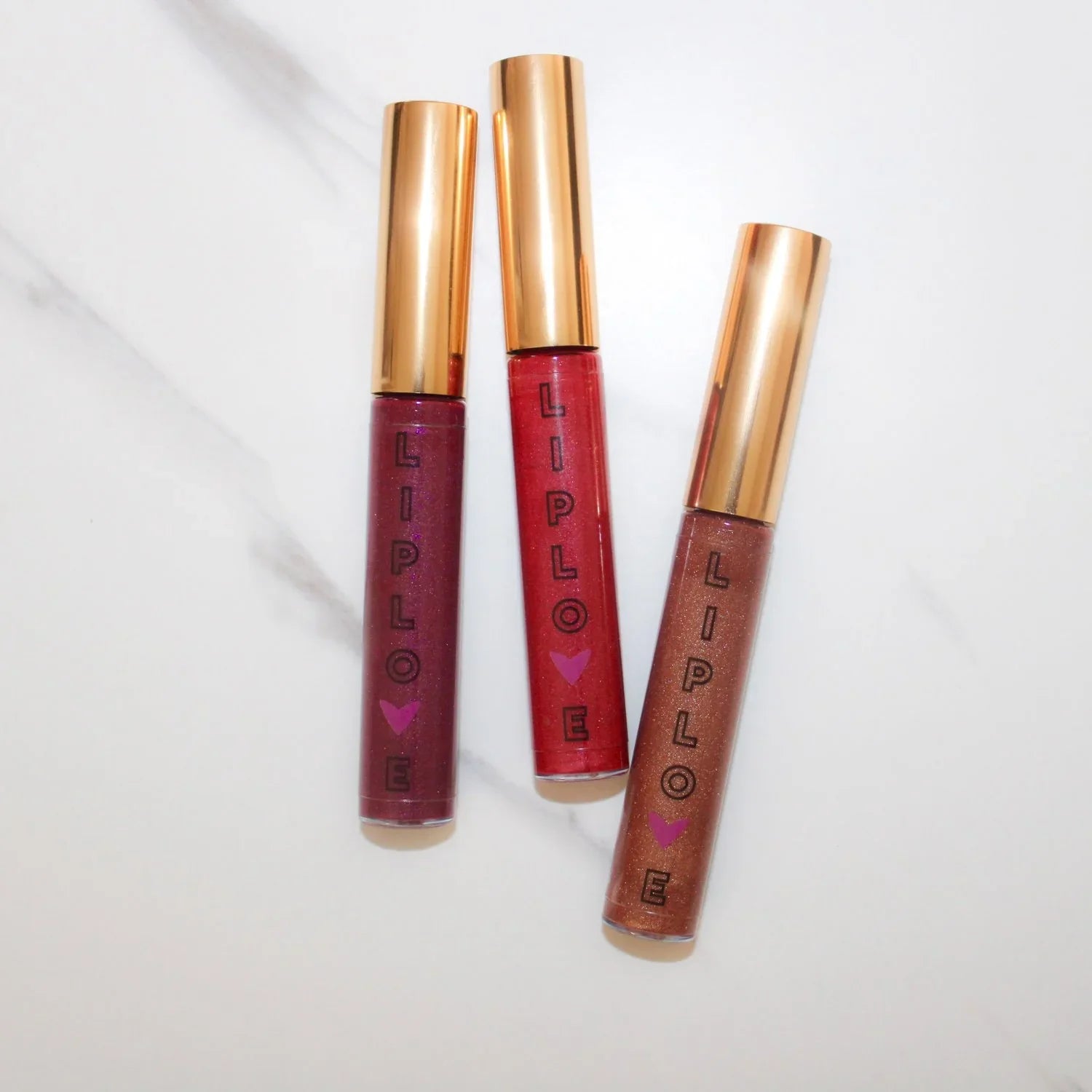 Three LIPLOVE lip gloss tubes with gold caps in red, mauve, and bronze on white marble