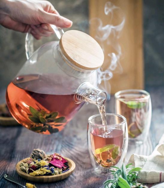 Hand pouring herbal tea from a glass teapot with wood lid into clear mugs, with dried flowers on table at Qu!rk Boutique.