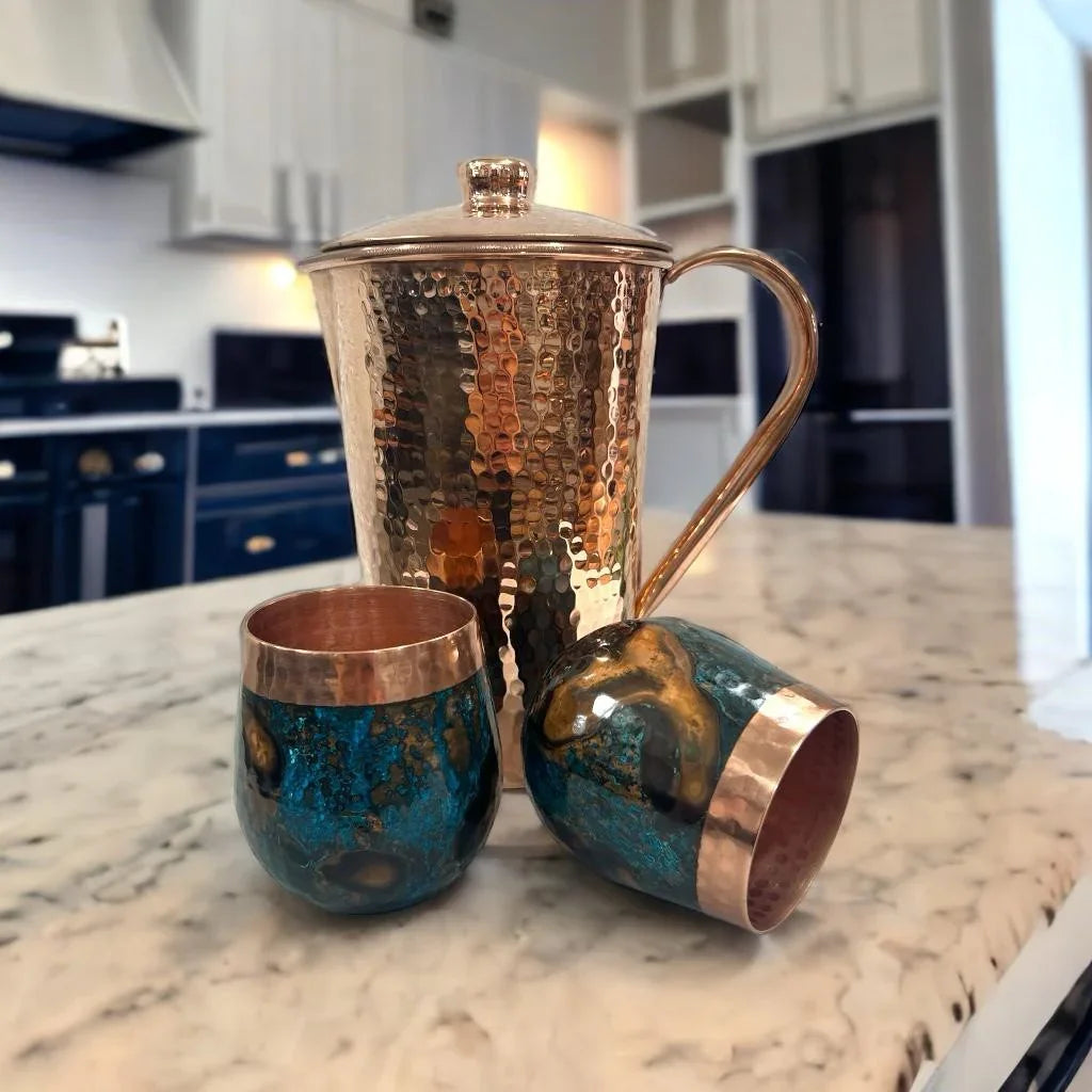 Hammered copper pitcher and two artistic blue and gold tumblers on marble kitchen counter.