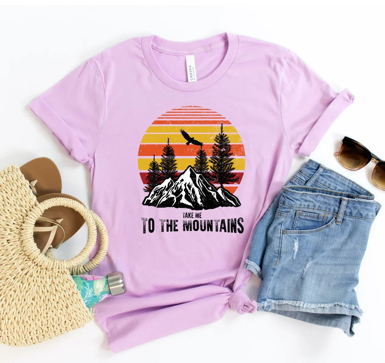 Lavender T-shirt with mountain and tree design, text 'Take me to the mountains', paired with denim shorts and accessories